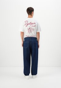White cotton t-shirt with “Dystopia Sounds” graphic in red, paired with loose-fitting dark blue denim pants and white sneakers.
