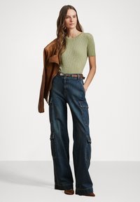 Ribbed light green short-sleeve top, high-waisted blue denim cargo pants with pockets, brown leather jacket draped over shoulder.