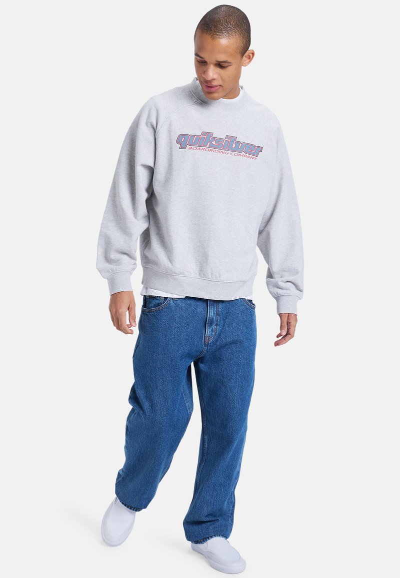Grå genser med lange ermer og ribbestrikkede mansjetter, med en rød Quiksilver-logo. Brukt med løse blå jeans og hvite joggesko.