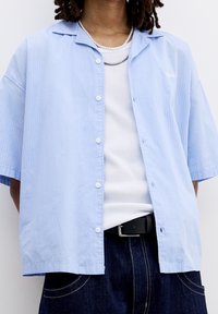 Camicia azzurra chiara a maniche corte con un motivo a righe verticali, tasche frontali e una vestibilità rilassata, indossata sopra una canotta bianca.