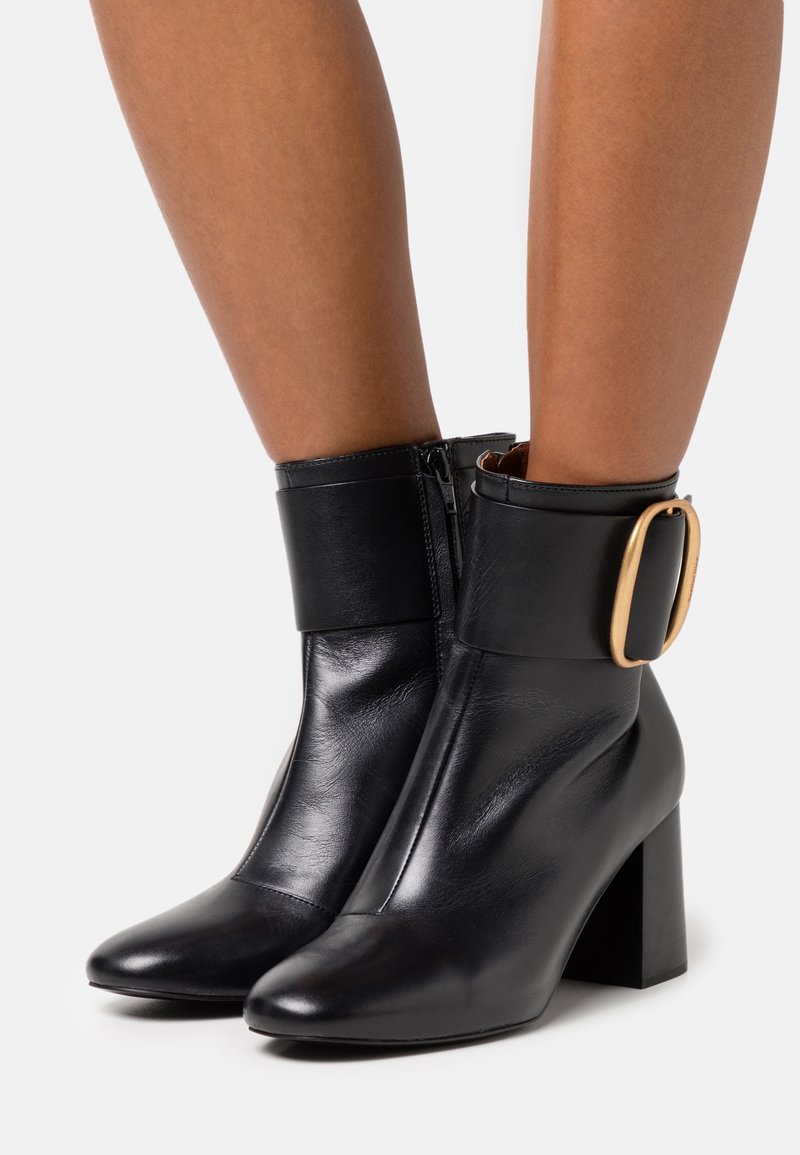 Černé kožené kotníkové boty se čtveratou patou, které mají velkou zlatou sponu na manžetě a boční zip pro zapínání.