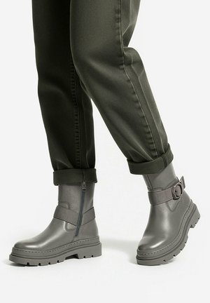 Nogi ubrany w podwinięte ciemnozielone spodnie i szare botki za kostkę z bocznymi zamkami, masywnymi podeszwami i paskami z klamrami.