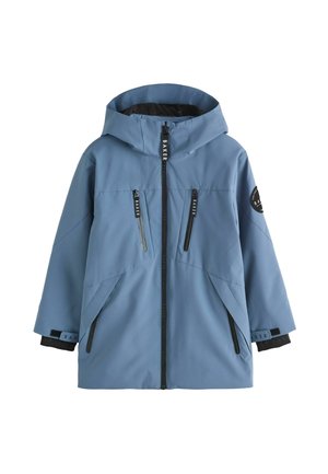 Junge steht mit den Händen in den Taschen, trägt eine blaue Kapuzenjacke, schwarze Cargohosen und weiße Turnschuhe vor einem einfarbigen Hintergrund.