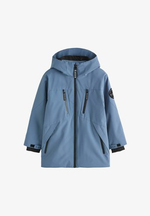 Junge steht mit den Händen in den Taschen, trägt eine blaue Kapuzenjacke, schwarze Cargohosen und weiße Turnschuhe vor einem einfarbigen Hintergrund.