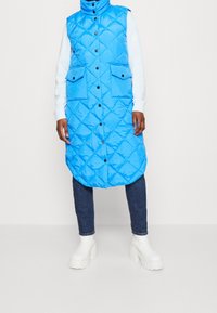 Blauwe gewatteerde bodywarmer met een hoge kraag, voorzien van diamantvormige stiknaden, twee voorzakken en een knoopsluiting. Draagt over een witte trui en een spijkerbroek.