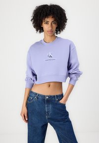 Levendula színű, rövidített kapucnis felső bordás mandzsettákkal és gallérral, melyen a "Calvin Klein Jeans" logó található. Laza fit stílusú sötét farmernadrággal viselve.