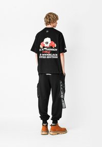 Camiseta negra de gran tamaño con un gráfico de calavera colorido en la espalda, combinada con pantalones cargo negros con bolsillos laterales y detalles en blanco. Botas marrones.