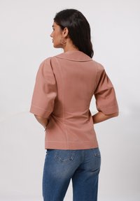Blusa strutturata rosa con maniche a palloncino corte, colletto a punta e dettagli in cucitura bianca. Indossata con jeans blu.