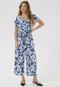 Jumpsuit blu e nero a fantasia con scollatura a V, maniche corte, vita cintrata e silhouette a gamba larga, abbinato a tacchi neri.
