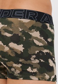 Boxers en camouflage vert et beige avec une taille élastique noire portant l'inscription "UNDER ARMOUR" en lettres grises. Texture de tissu douce.
