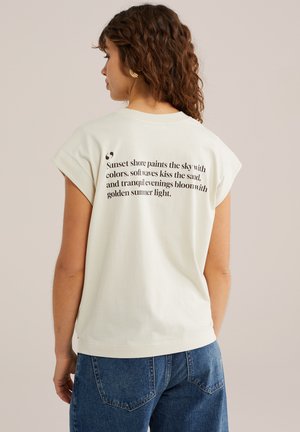 Žena s kučeravými vlasmi nosí biele tielko s čiernym poetickým textom na chrbte, v kombinácii s modrými rifľami, neutrálnym pozadím.