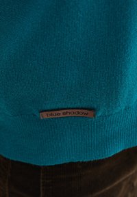 Turkusowa dziana tkanina z małą brązową skórzaną metką przyszytą, z wytłoczonym napisem "blue shadow", nad ciemno brązowym materiałem kordurowym.