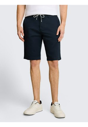 Mann trägt marineblaue Shorts mit Kordelzug und weiße Sneakers, steht vor schlichtem hellem Hintergrund.