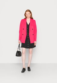 Blazer duplo de botão rosa brilhante sobre um vestido preto com detalhes em botão. Uma mala preta estilo balde e sapatos de salto bloco pretos completam o look.
