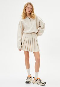 Suéter grande beige con cuello de media cremallera, combinado con una mini falda plisada beige. Lleva zapatillas deportivas y calcetines blancos.