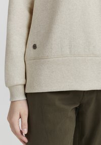 Beige genser laget av mykt stoff, med ribbestrikket mansjett og en liten metall-logo detalj nær kanten. Sammen med olivengrønne bukser.