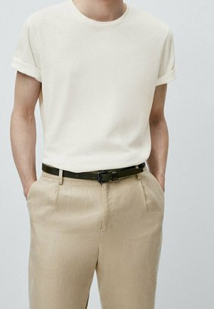 Homme portant un t-shirt uni écru à manches courtes, rentré dans un pantalon beige avec une ceinture noire, mains dans les poches.