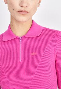 Rosa geripptes Polo-Shirt mit einem Viertelreißverschluss, flachem Kragen und dezentem Lacoste-Logo auf der Brust. Glatte Textur.
