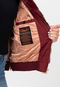 Giacca bomber color bordeaux con colletto in pelliccia ecologica; fodera interna in satin con etichetta nera e testo arancione; chiusura con zip dorata e polsini a coste.