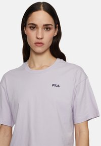 Violetti puuvillapaita, jossa on pyöreä kaula-aukko ja lyhyet hihat. Edessä vasemmalla rinnassa on pieni musta FILA-logo.
