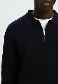 Marineblauer Strickpullover mit klassischem Kragen und kurzem Reißverschluss, mit strukturierter Stoffoberfläche, sichtbaren Nähten und kontrastierendem Innenfutter.