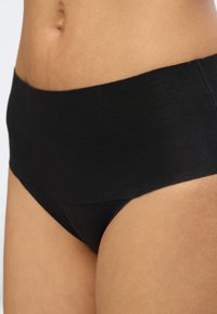 Gros plan sur la taille et les hanches d'une personne portant une culotte noire taille haute sur un fond uni.