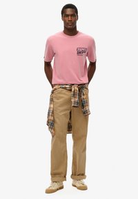 Hombre con camiseta rosa de Superdry, pantalones beige, zapatillas beige, con una camisa de cuadros amarilla atada a la cintura, de pie contra un fondo blanco.