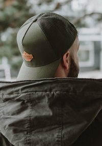 Cappellino da baseball verde oliva con retro in rete, caratterizzato da una patch in pelle marrone con il logo del marchio, indossato da una persona con una giacca scura.