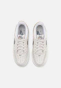 Sněrovací tenisky Nike Air Force 1 v světle béžové kůži s perforovanou špičkou, bílými tkaničkami, šedými akcenty a polstrovanou podešví.