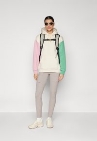 Hettegenser med pastellrosa, grønn og kremfargede blokker, frontlomme, kombinert med lysegrå cargo-bukser og beige joggesko. Svart ransel med hoftebelte.