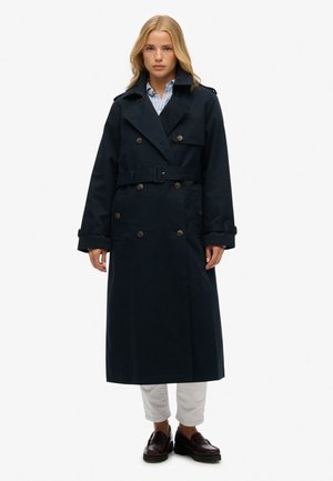 Lange marineblauwe trenchcoat met dubbelrijige knopen, riem, grote kraag en manchetten. Gemaakt over een lichtblauw gestreept overhemd en witte broek.