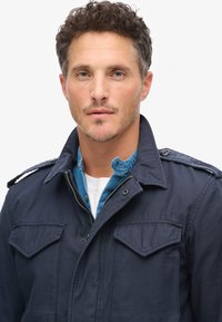 Mørkeblå jakke med lynlås og trykknaplukninger, skulderlapper og to brystlommer over en lagdelt lyseblå skjorte. Tekstureret stof.