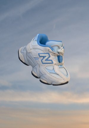 Chaussure de sport blanche et bleu clair avec une semelle épaisse flottant devant un ciel nuageux.