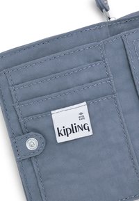 Šedá peněženka chránící RFID, která má více přihrádek na karty, zapínání na cvok a texturovaný látkový povrch. Viditelná visačka s logem Kipling.