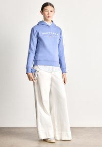 Sudadera con capucha azul claro con logo blanco, combinada con una camiseta blanca de rayas debajo. Pantalones anchos blancos y suelas plateadas tipo slip-on.