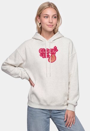 Ung kvinde iført hvid hættetrøje med pink og orange grafisk tekst og lyseblå jeans står med den ene hånd på hoften.