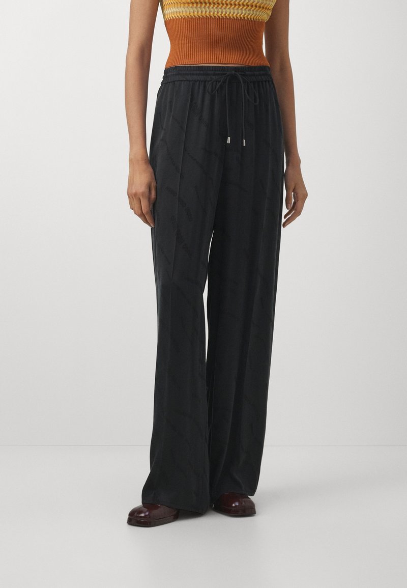 Personne portant un pantalon noir ample avec une taille à cordon, associé à un haut tricoté sans manches orange et jaune et des chaussures marron foncé.