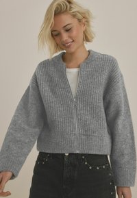 Grå stickad cardigan med ribbad textur, rundad halsringning och hel dragkedja framtill. Bärs över en vit topp och kombineras med mörka jeans.