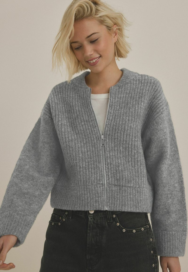 Grå stickad cardigan med ribbad textur, rundad halsringning och hel dragkedja framtill. Bärs över en vit topp och kombineras med mörka jeans.