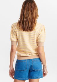 Kvinde med bølget brunt hår iført en pink og gul ternet kortærmet sweater og blå denimshorts, stående med ryggen til.