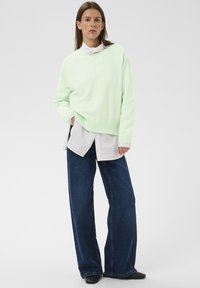 Naine kannab helerohelist kampsunit valge särgi üle, laia säärega siniseid pükse ja musti kingi, seistes valge ühevärvilise tausta ees.