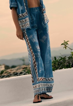 Person trägt blaue gemusterte weite Hose und passendes offenes Hemd, steht draußen auf einer weißen Brüstung mit Pflanzen und Horizont im Hintergrund.