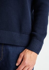 Jūras zila džemperis ar teksturētu adījuma rakstu, ar ribotiem piedurknēm un apakšmalu, apvienots ar džinsu biksēm. Tuvplāns ar roku un auduma detaļām.