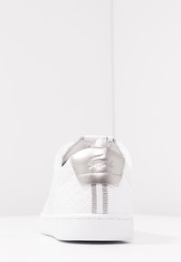 Zapatilla blanca con parte superior texturizada, lengüeta plateada en el talón y suela de goma blanca. Presenta una lengüeta de agarre y una marca detallada en el talón.