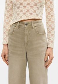 Μπεζ παντελόνι ευρείας γραμμής με λεία υφή, πέντε τσέπες και κλείσιμο με μεταλλικό κουμπί. Συνδυασμένο με ένα κρεμ δαντελένιο τοπ που είναι cropped.