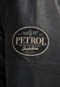 Zwarte leren jas met een ronde, geborduurde patch met het "PETROL"-logo, vleugels en tekst in wit en crème aan de linkerkant.