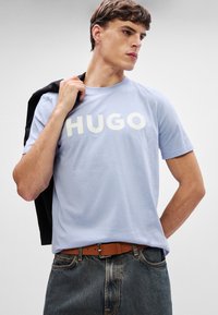 Ανοιχτό μπλε βαμβακερό t-shirt με λευκό λογότυπο "HUGO", στρογγυλή λαιμόκοψη, κοντά μανίκια, σε συνδυασμό με γκρι τζιν παντελόνι και καφέ ζώνη.