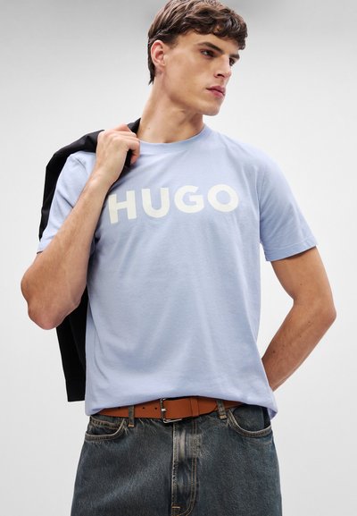 Светлосиня памучна тениска с бял логотип "HUGO", с кръгло деколте и къси ръкави, съчетана с сиви дънки и кафяв колан.