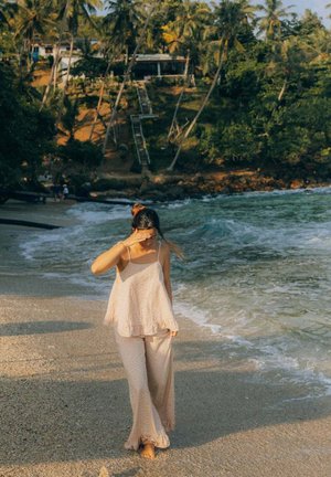 Kobieta w jasnym piżamowym zestawie spaceruje boso po piaszczystej plaży przy oceanie, osłaniając oczy przed światłem słonecznym, w tle palmy i domy.