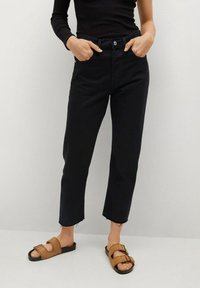 Pantalones vaqueros cortos de mezclilla negra con una cintura ajustada, bolsillos frontales y dobladillo deshilachado. Combinados con sandalias marrones de tipo deslizable con hebillas.